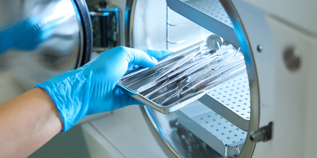 Gloved hand holding a metal tray with dental tools in a sterilization machine.